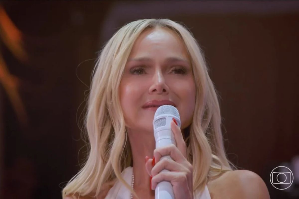 Apresentadora Angélica segura um microfone branco e canta em um palco com iluminação dramática. Ela tem cabelos loiros e usa…