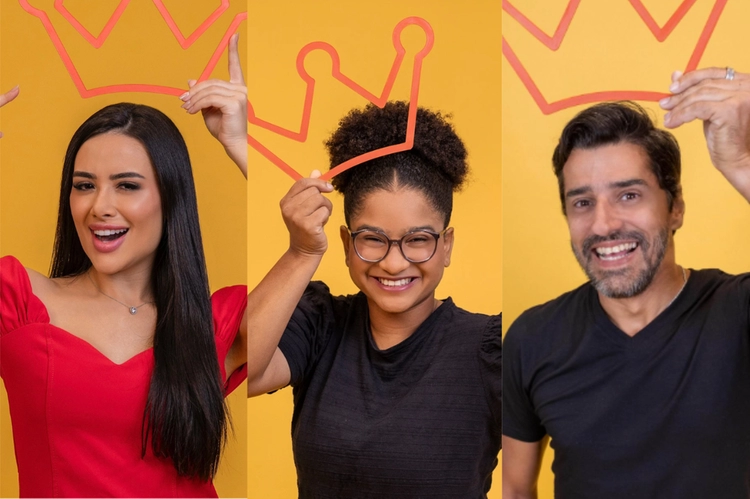Três pessoas sorrindo e segurando coroas de papelão sobre a cabeça: uma mulher de vestido vermelho, uma jovem de óculos e ca…