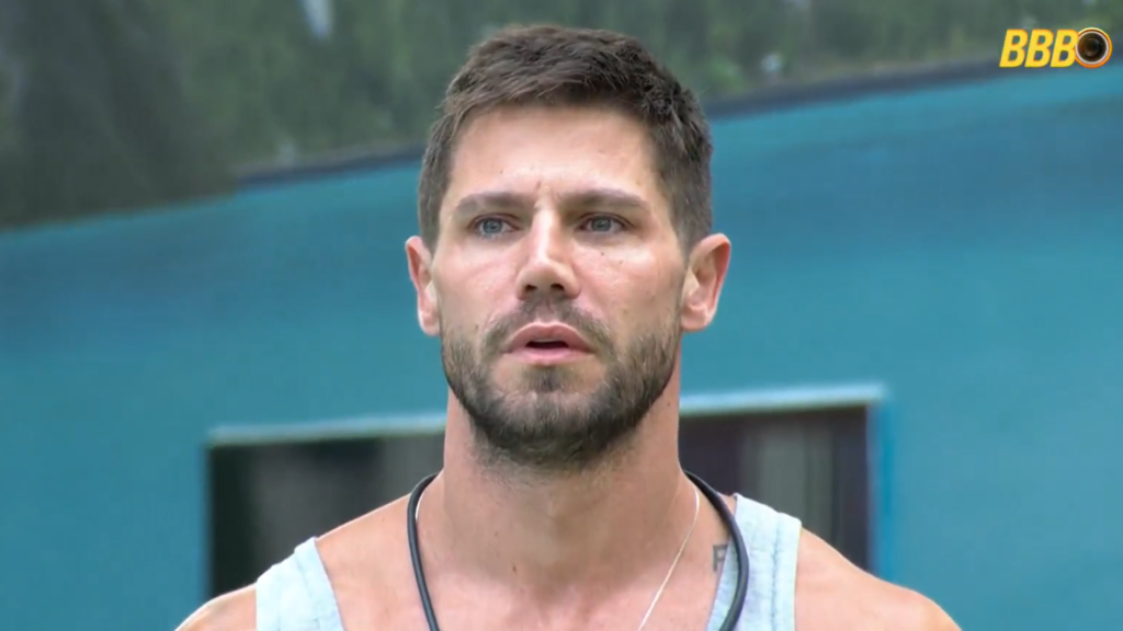 Homem com barba e cabelo curto, vestindo regata cinza, com expressão séria. Logo do BBB ao fundo.