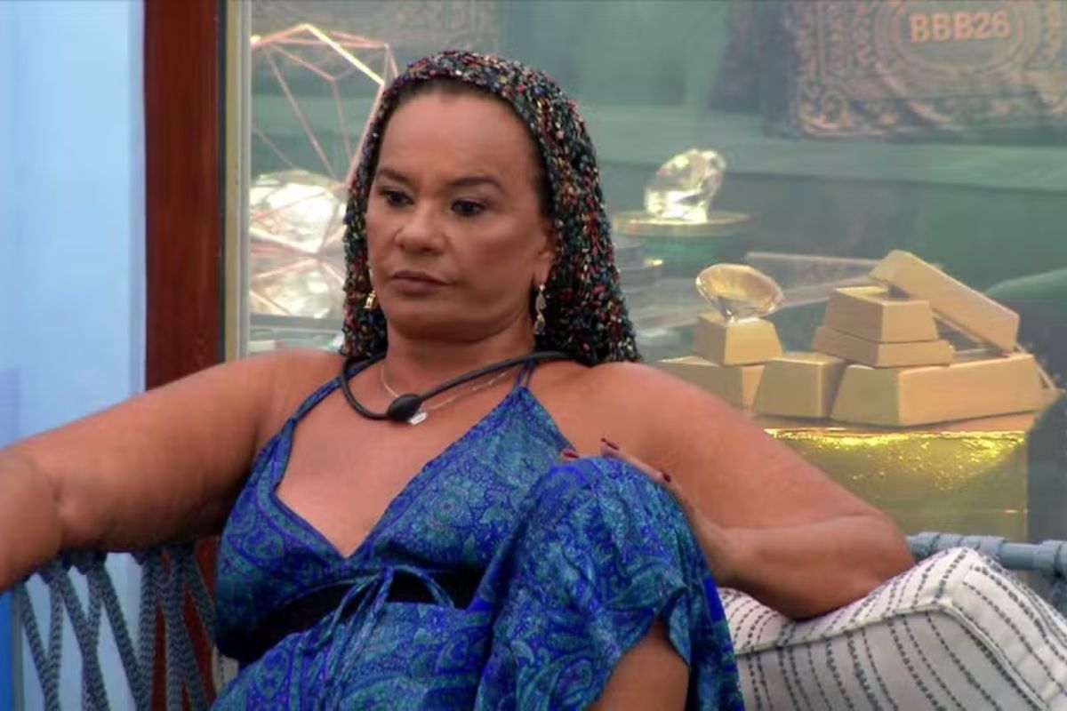 Participante do BBB 26, Dona Geralda, usa um vestido azul estampado e um lenço colorido na cabeça, sentada em uma poltrona.