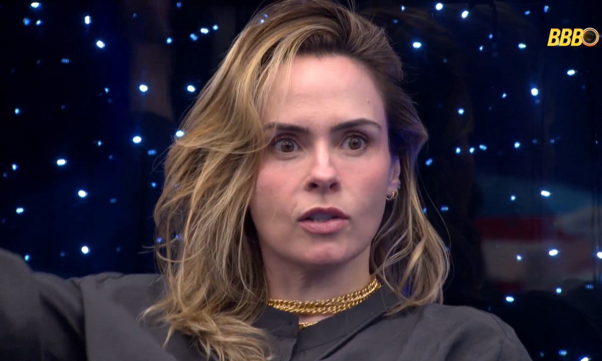 Apresentadora do BBB, Tadeu Schmidt, em close-up com fundo escuro e luzes azuis, usando colar dourado.