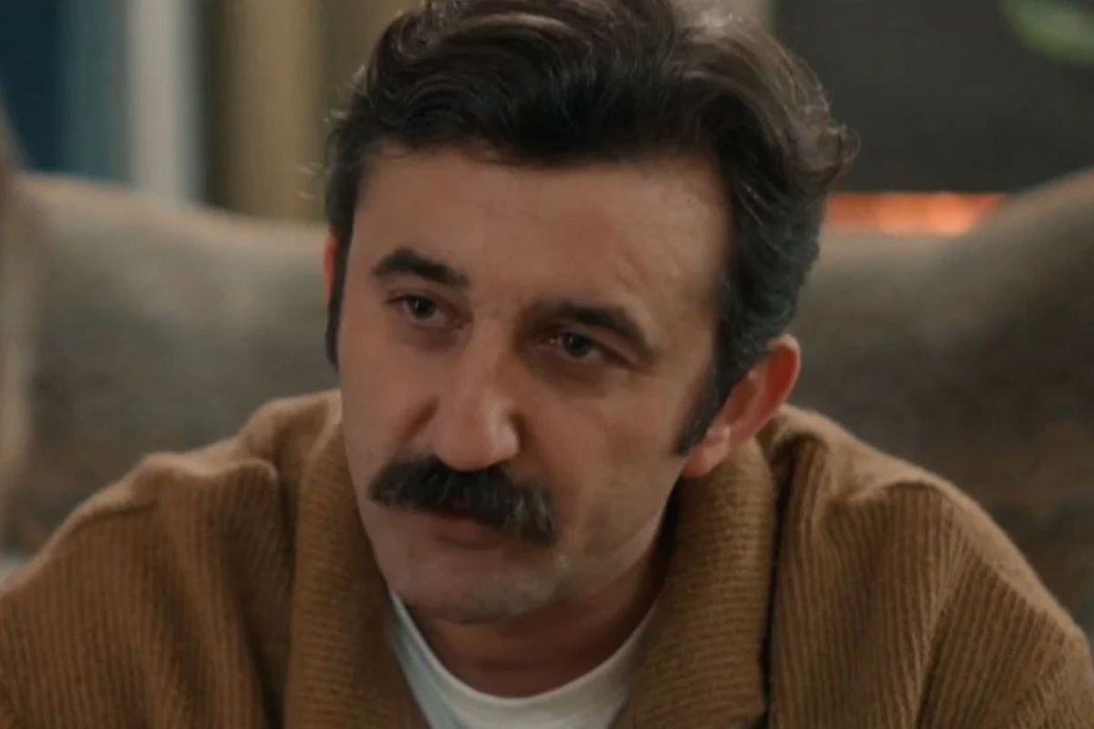 Homem com bigode proeminente e cabelo escuro penteado para trás, vestindo um suéter marrom.