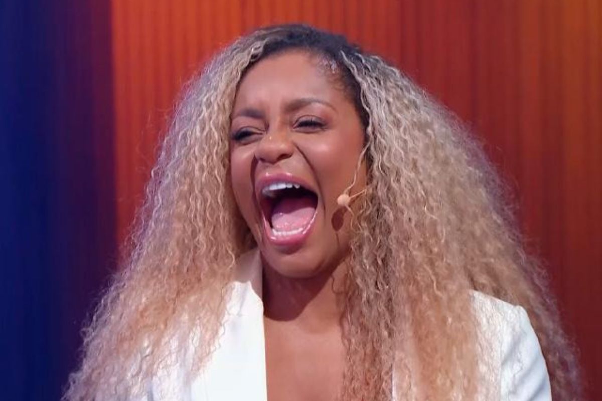 A mulher com cabelo cacheado loiro e branco, vestindo um blazer branco, grita com a boca aberta em um programa de TV.