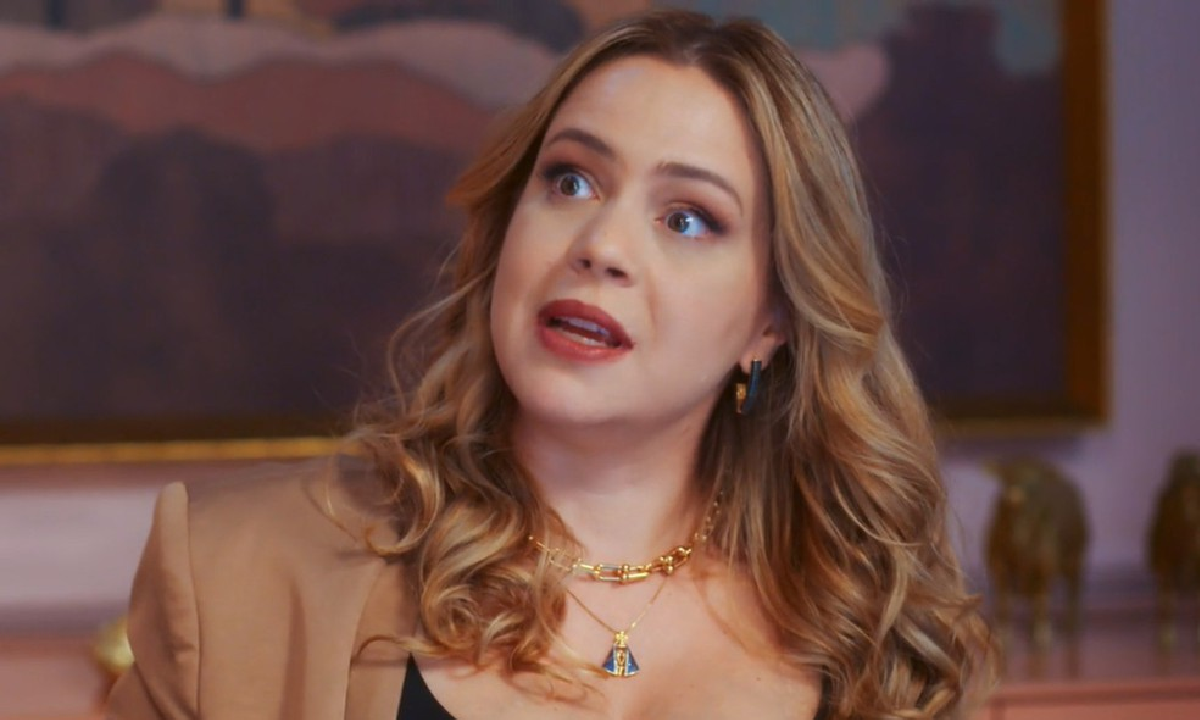 Atriz Agatha Moreira em cena de novela, com cabelo loiro ondulado e maquiagem marcante, usando blazer bege e colares dourado…